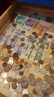 Drawer containing a variety of paper bills from multiple countries and numerous assorted coins scattered on the surface.
