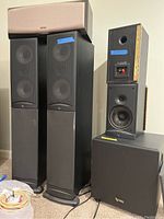 Photo showing six different Infinity speaker components including two tall floor speakers, one center speaker, two smaller speakers (one without grille), and one heavy subwoofer with visible wiring and ports.