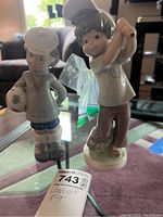 Two Lladro figurines shown side-by-side on a glass table, one holding a soccer ball and wearing a beret, the other a golfer swinging with a cap.