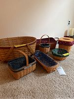 Overall group of six woven Longaberger baskets and one tissue holder with lid, displayed on carpet, showing various sizes and cloth/plastic liners.