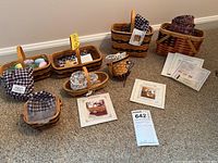 Photo showing seven Longaberger miniature baskets arranged on carpeted floor with various fabric liners and decorative contents, including Easter eggs and hay filler. Several information cards and tags are visible on or near baskets.