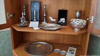 Wide view of upper cabinet shelf showing pewter candlesticks, metallic horse figurine, a decorative bowl, and a tray along with small plates and box.