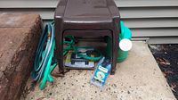 2 brown plastic side tables, garden hoses, Sigma Metal Spike-A-Lite watering timer in packaging, and green watering can arranged outside on concrete.