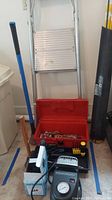 Photo of metal step stool, two blue-handled sledgehammers, red toolbox partially open showing various tools, black air pump, and other items arranged on garage floor.