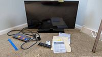 Front view of Samsung TV on stand with remote, power strip, cables, and manuals beside it on carpeted floor.
