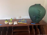 Wide view of two frog figurines, ceramic tile, and green glass vase on wooden shelf
