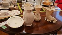 Four Belleek porcelain pieces on wooden table including three vases and one trinket bowl, showing overall lot contents.