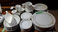 Photo showing the complete set including stacked plates, cups, saucers, and gravy boat, with green and gold vine floral pattern on white china.