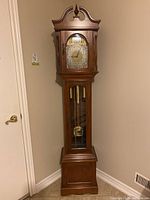 Front view of a tall granddaughter clock showing wooden case, clock face with brass pendulum and weights visible through glass panel.