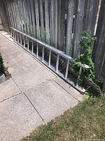Full side view of aluminum extension ladder leaning against wooden fence on sidewalk showing entire length.