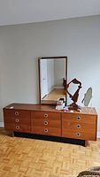 Front view of nine-drawer teak veneer dresser with oval metal pulls and matching mirror leaning against wall