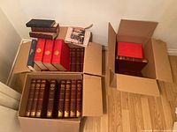Photo of three open boxes containing multiple hardcover books including red, dark blue, and brown leather-bound volumes arranged inside boxes on a wooden floor.