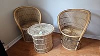 Pair of vintage wicker arm chairs with rounded backs and matching wicker side table with glass top on hardwood floor against white wall.