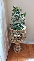 Full view of wicker plant stand with faux ivy plant inside located in corner against white wall and hardwood floor