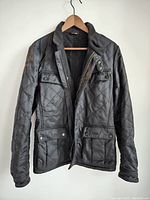Front view of the black quilted Barbour men's jacket hanging on a wooden hanger, showing zip and button flap pockets, and collar details.