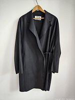Front view of the black Acne Studios coat hanging on a wooden hanger showing belted waist and lapel collar.