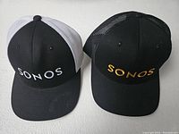 Two Sonos branded trucker hats with mesh backs, one with white mesh and white embroidery, the other with black mesh and gold embroidery.