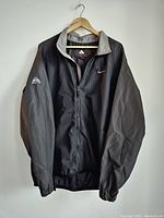 Front view of Nike ACG men's jacket on hanger showing black body and gray sleeves, with Nike swoosh and ACG logo embroidery on sleeve.
