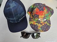 Top-down image showing the pair of black framed Oakley polarized sunglasses and two Oakley hats - one tropical print and one blue camo.