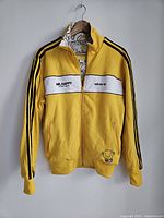 Front view of yellow Adidas jacket with Mr Happy and Adidas logos, black stripes on sleeves, zipper closed.
