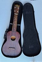 Full view of the Aria ukulele open in its hard case showing the wood grain and headstock logo.