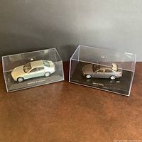 Two diecast car models in display cases on a brown surface, showing both Maserati and BMW cars from an angled front view.