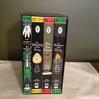 Front view of the boxed set featuring spines of four books: The Hobbit, The Fellowship of the Ring, The Two Towers, and The Return of the King. The slipcase is decorated with green, red, and yellow Tolkien-themed symbols and designs.