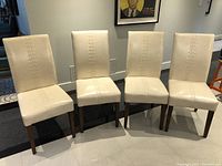 Four cream colored faux leather dining chairs with wooden legs and vertical stitched detail aligned side by side.