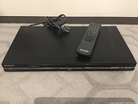 Photo showing the Sony DVD player on a carpeted floor with the power cord and remote control placed on top.