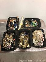Full lot showing five trays with assorted costume jewelry pieces including necklaces, earrings, brooches, pins and charms mixed together