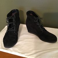 Pair of black suede lace-up booties with wedge heels shown on white cloth, displaying front and side views.