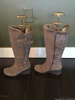 Pair of light brown mid-calf suede leather boots with fur lining, side zipper and buckle strap detail shown side by side on dark floor against a gray wall.