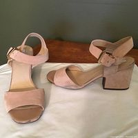 Front and side view of the pair of beige suede heeled sandals with ankle strap buckle and block heel, placed on white surface against dark background.