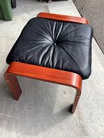 Side angle showing black leather cushioned top with a central button, attached to curved teak wood frame with brass screws.