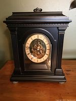Front view of the black Bombay Company mantel clock showing the clock face with Roman numerals and gold-tone mechanical interior visible through the glass.