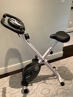 Front angle of the white folding exercise bike with black padded seat and handlebars, showing frame and pedals.