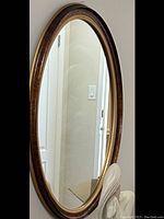 Photo of an oval wall mirror with a brown wood frame and a gold inner border. The mirror is hung on a light-colored wall with part of a statue visible in the lower right corner, not included in sale.