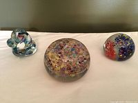 Photo showing three glass paperweights placed on a white surface against a plain background. From left to right: a swirl-shaped colored glass paperweight, a large round millefiori paperweight, and a round paperweight with colorful floral inclusions.