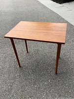 Photo shows a small teak table with rectangular top and tapered legs standing on a gray paved surface outdoors. Visible wood grain and some scratches on the top surface.