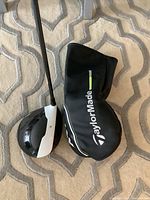 Photo of black and white TaylorMade M1 driver clubhead with included black TaylorMade head cover on carpet.