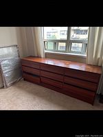Full view of the long teak wood dresser positioned against a wall with windows, showing the 9 drawers in three columns, top surface, and overall condition.