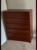 Teak highboy dresser with four drawers, showing front view against a white wall on carpeted floor.