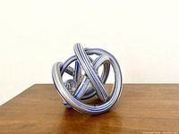 Front angled view of blue art glass knot sculpture showing intertwined loops and embedded gold glitter inside transparent blue glass.