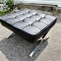 Full side angle of black leather tufted ottoman with chrome legs on pavement.