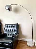 Full view of lamp showing arched chrome rod, hemispherical shade, round marble base beside armchair