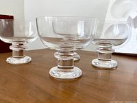 Front view showing all four clear glass pedestal glasses arranged in a group on a wooden surface, highlighting their shape and design.