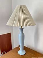 Full view of pale blue Mid-Century Modern lamp with beige pleated conical lampshade on wooden surface.