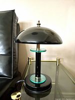 Front side view of a black mid-century modern mushroom-style table lamp with green glass discs on the stem and base, placed on a glass tabletop.