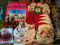 Top-down view showing assorted Christmas placemats, napkins, hot pads, and plastic tablecloth with various holiday designs including Santa faces and winter scenes.