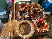 Photo of five straw wreaths (some decorated with fall-themed embellishments) and four straw brooms with wooden handles laid out on a surface.